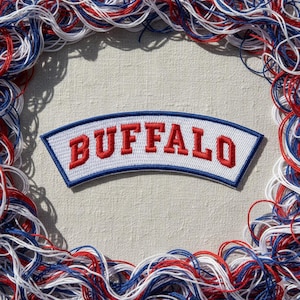 May include: Embroidered patch with the word "BUFFALO" in red letters on a white background, framed by a blue border. The patch is surrounded by red, white, and blue threads, creating a decorative border. The patch is centered on a neutral-toned fabric.