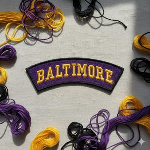 May include: A purple patch with the word "BALTIMORE" embroidered in yellow. The patch is surrounded by yellow, purple, and black embroidery floss on a light-colored fabric background.