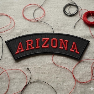 May include: A black embroidered patch with the word "ARIZONA" in red, arched across the top. Red and black thread spools and loose threads are scattered around the patch on a neutral-colored fabric.