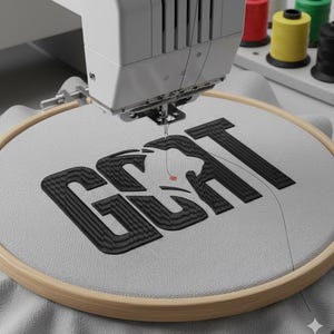 May include: An embroidery machine stitching the word "GOAT" in black thread onto a light gray fabric stretched within a wooden hoop. Spools of colorful thread are visible in the background. The machine is in operation, creating a textured design.