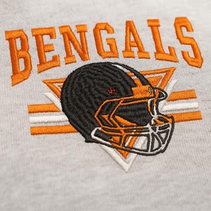 Peut inclure: Broderie avec le mot "BENGALS" en orange au-dessus d'un casque de football américain noir et orange. Le casque est orné de détails blancs et orange, sur un fond en tissu gris clair. Le motif comprend un triangle blanc et des rayures orange.