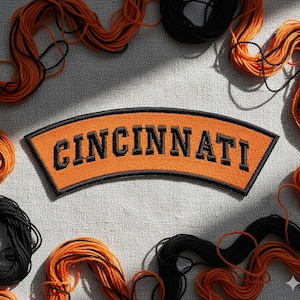 Puede incluir: Un parche naranja con la palabra "CINCINNATI" bordada en negro, arqueada en la parte superior. Hilos negros y naranjas rodean el parche, lo que sugiere un proyecto de artesanía o bricolaje.