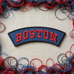May include: A blue embroidered patch with the word "BOSTON" in red, arched across the center. The patch is framed by a black border and surrounded by red, blue, and black threads, creating a decorative border.