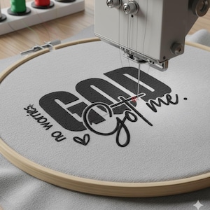 May include: An embroidery machine stitching the phrase "God Got me" in black thread onto a light gray fabric stretched within a wooden hoop. The words "no worries" and a heart are also embroidered.