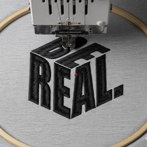 Puede incluir: Una máquina de bordar está bordando las palabras "BE REAL" en hilo negro sobre tela gris claro. El texto está en un estilo de letras de bloque 3D, dentro de un bastidor de bordado de madera.