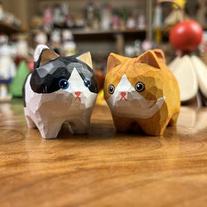 May include: Two wooden cat figurines. One is black and white, the other is orange and white. Both have large blue eyes and red noses. The cats are carved with a geometric style and sit on a wooden surface.