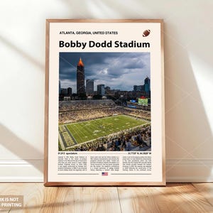 May include: A framed print of Bobby Dodd Stadium in Atlanta, Georgia. The image shows the stadium's field, filled with spectators, with the city skyline in the background. Text includes the stadium name and location.