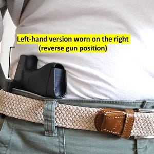 Op de afbeelding: Zwarte holster gedragen op een beige gevlochten riem met een bruine leren gesp. De holster bevindt zich aan de rechterkant, zoals aangegeven door de tekst "Left-hand version worn on the right (reverse gun position)". De persoon draagt een wit shirt en een groene broek.