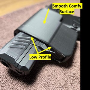 Op de afbeelding: Close-up van een zwart object met een glad, comfortabel oppervlak en een laag profiel ontwerp. Het object lijkt gemaakt te zijn van een mat materiaal. De tekst "Smooth Comfy Surface" en "Low Profile" zijn zichtbaar.