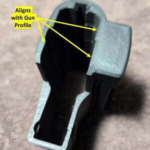 May include: A gray 3D-printed object with the text "Aligns with Gun Profile" and yellow arrows pointing to the edges. The object has a hollow interior and a textured surface, designed to fit a gun profile.