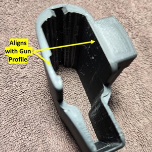 May include: A gray and black 3D-printed object with the text "Aligns with Gun Profile." The object has a curved shape with a rectangular opening and a textured surface. The object appears to be a holder or a component.