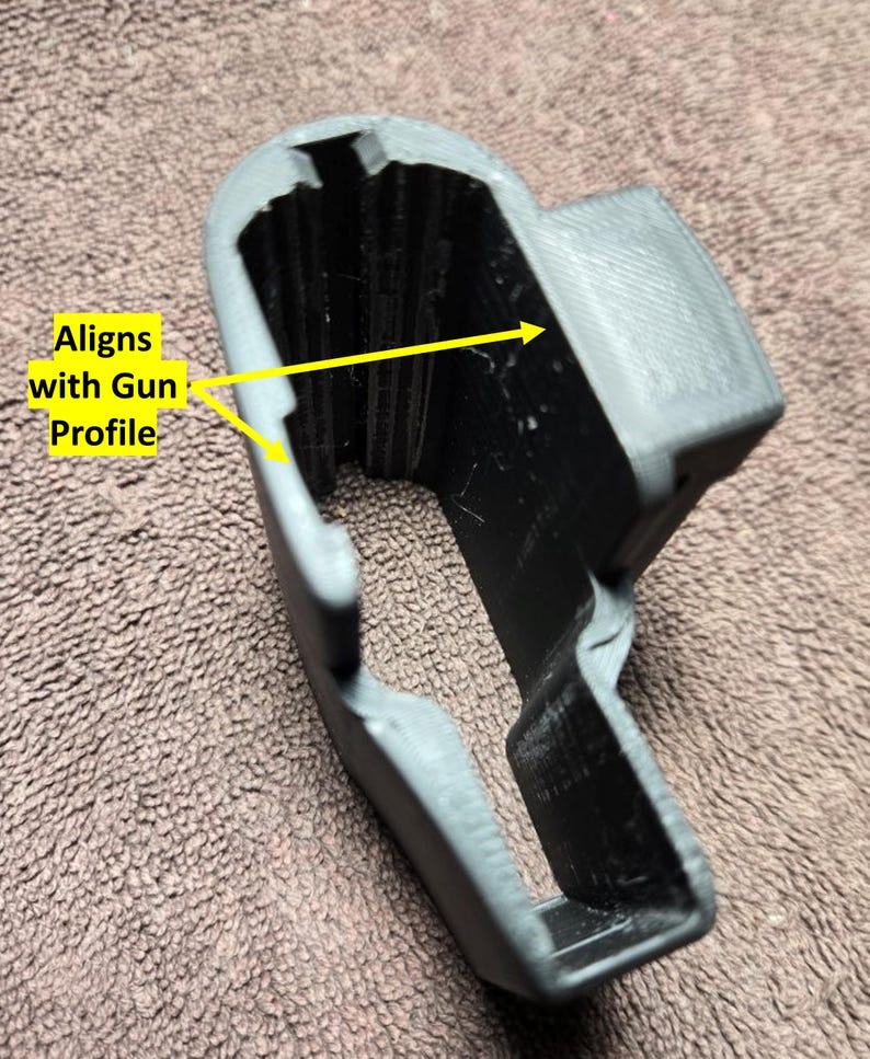 May include: A gray and black 3D-printed object with the text "Aligns with Gun Profile." The object has a curved shape with a rectangular opening and a textured surface. The object appears to be a holder or a component.