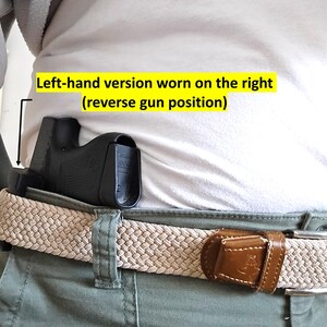 May include: A black holster is worn on the right side, attached to a beige woven belt with a brown leather buckle. The text in the image reads: "Left-hand version worn on the right (reverse gun position)". The person is wearing a white shirt and green pants.