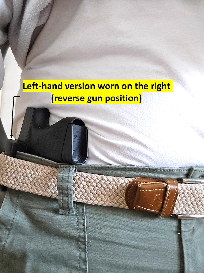 May include: A black holster is worn on the right side, attached to a beige woven belt with a brown leather buckle. The text in the image reads: "Left-hand version worn on the right (reverse gun position)". The person is wearing a white shirt and green pants.