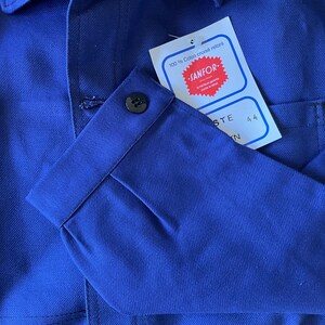 Op de afbeelding: Een blauw werkshirt met een geknoopte manchet en een label met de tekst "100% Coton crois&eacute; retors" en "SANFOR." Het label heeft ook de maat "44" erop gedrukt.
