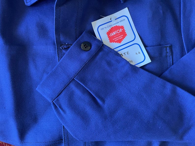 Op de afbeelding: Een blauw werkshirt met een geknoopte manchet en een label met de tekst "100% Coton crois&eacute; retors" en "SANFOR." Het label heeft ook de maat "44" erop gedrukt.