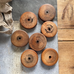 May include: A collection of six circular wooden spools, each featuring a central hole and a natural brown wood grain finish with varying dark markings. The spools are arranged in a cluster, showcasing their smooth, flat surfaces and rounded edges.