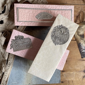 Puede incluir: Una colección de artículos vintage con temática de bautizo, que incluye cajas rosas y un sobre color crema, todos adornados con la palabra "Bapteme" en letras plateadas. También hay un retrato de una mujer con un vestido azul.