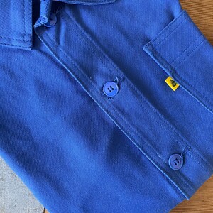 Op de afbeelding: Een blauw werkshirt met twee knoppen en een gele patch op de zak.