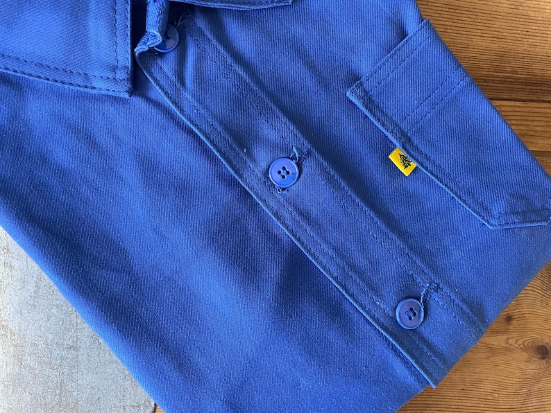 Op de afbeelding: Een blauw werkshirt met twee knoppen en een gele patch op de zak.