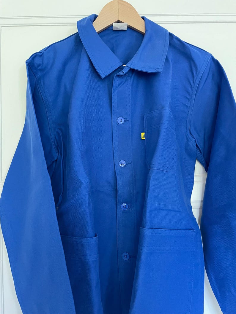 Op de afbeelding: Een blauwe werkjas met een knoopsluiting en twee grote zakken. De jas heeft een geel label aan de voorkant.