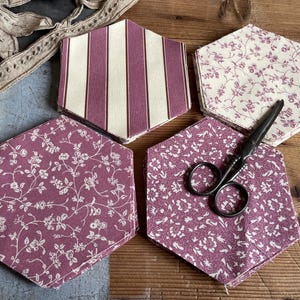 May include: Hexagonal fabric pieces with floral and striped patterns in purple and cream. A pair of black scissors sits on one of the floral pieces. The items are arranged on a wooden surface. The fabric pieces are likely for crafts.