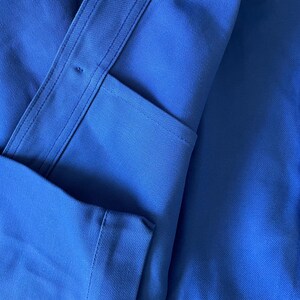 Op de afbeelding: Een close-up van een blauw werkshirt met een knoopzak en een knoopsluiting aan de rechterkant.