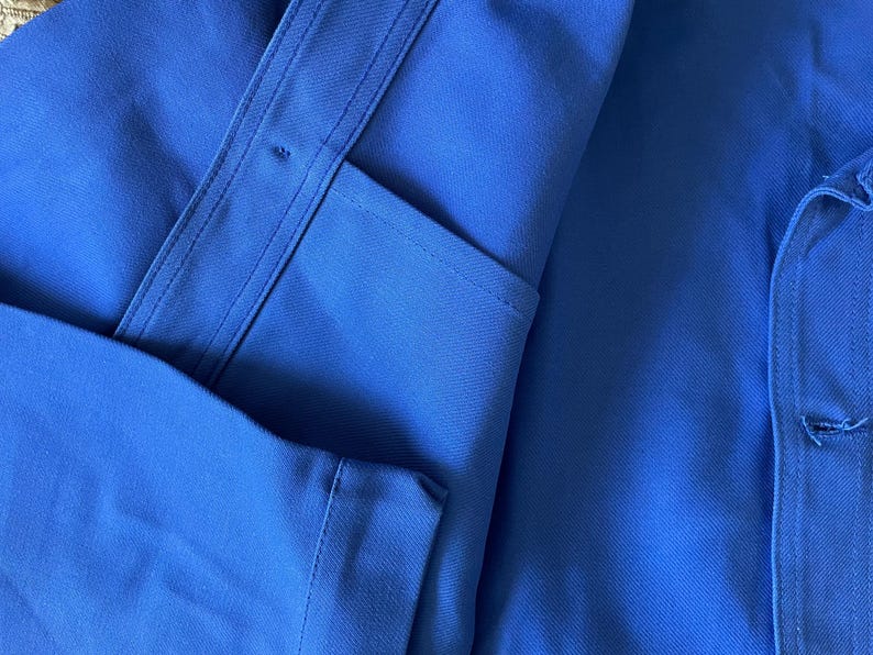 Op de afbeelding: Een close-up van een blauw werkshirt met een knoopzak en een knoopsluiting aan de rechterkant.