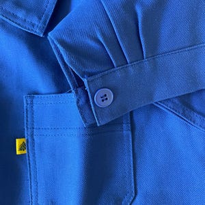 Op de afbeelding: Een close-up van een blauw werkshirt met een geel logo op de zak en een knoop op de mouw.