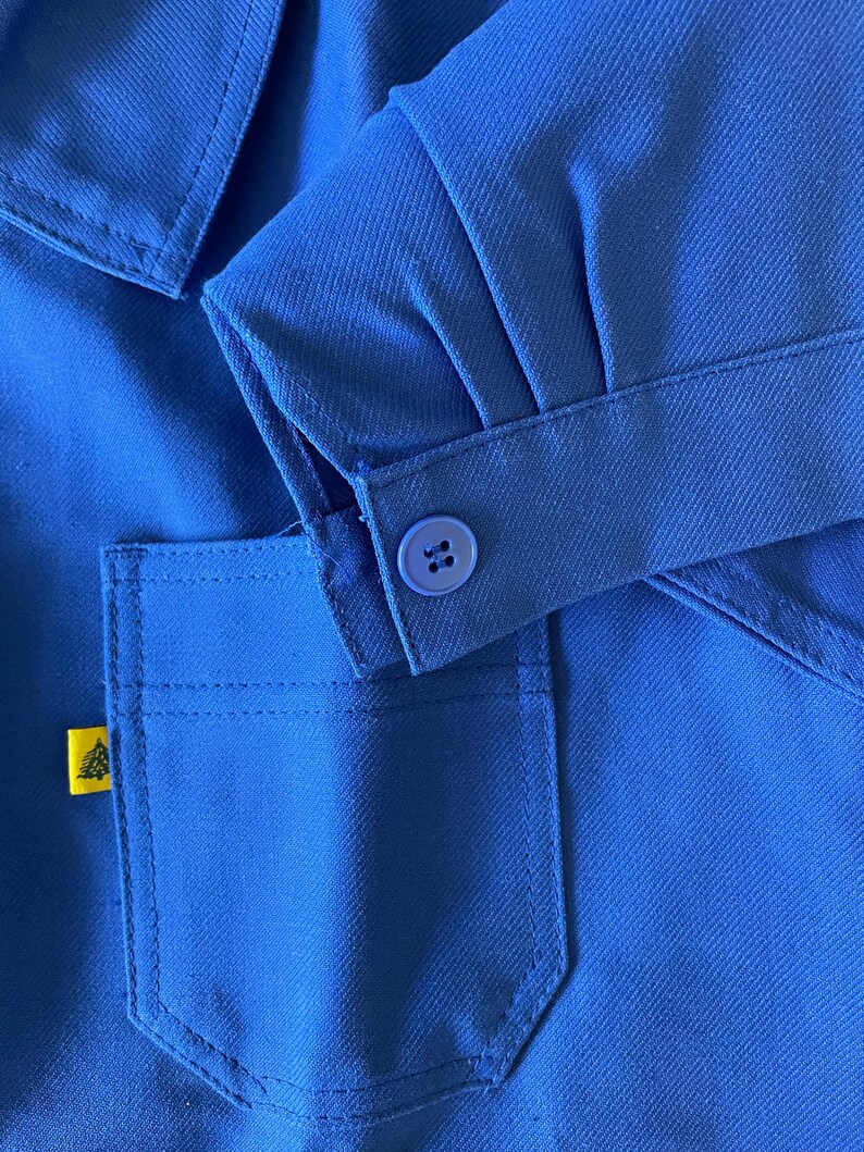 Op de afbeelding: Een close-up van een blauw werkshirt met een geel logo op de zak en een knoop op de mouw.