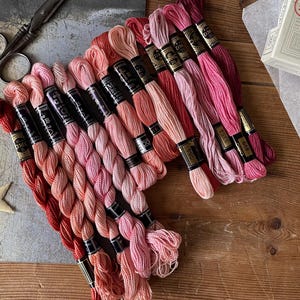 Pode incluir: Uma coleção de meadas de linha de bordar rosa e vermelha, dispostas em um gradiente de cores. Uma tesoura vintage e uma decoração em forma de estrela também são visíveis. Uma caixa de linha de bordar DMC está ao fundo.