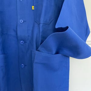 Op de afbeelding: Een blauwe werkjas met een geel logo op de borst en twee grote zakken aan de voorkant.