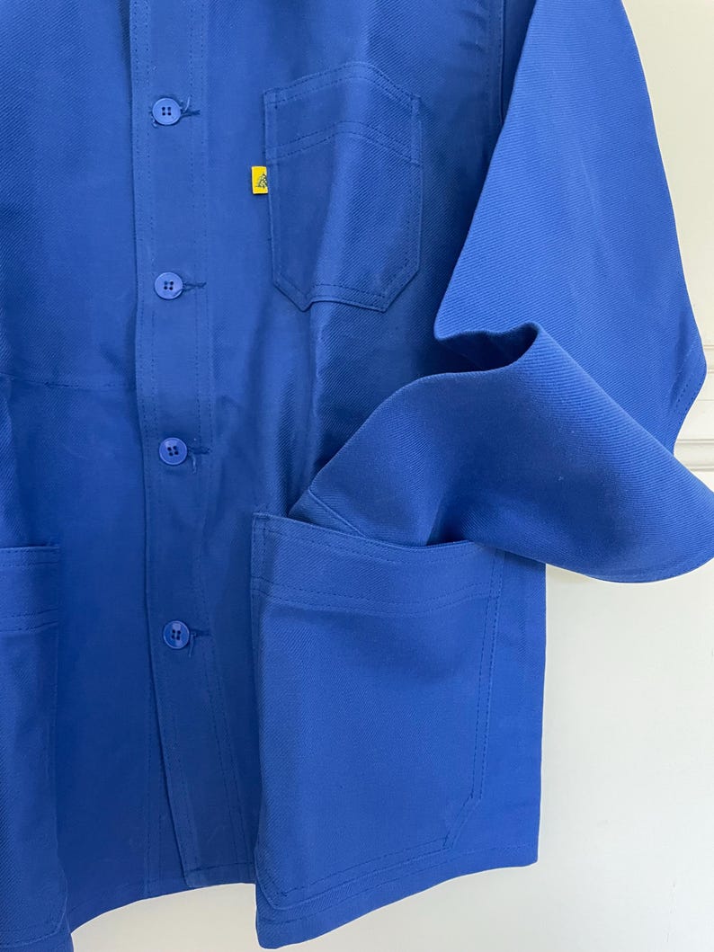 Op de afbeelding: Een blauwe werkjas met een geel logo op de borst en twee grote zakken aan de voorkant.