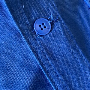 Op de afbeelding: Close-up van een blauwe knoop die op een blauw stoffen shirt is genaaid. De knoop heeft vier gaten.