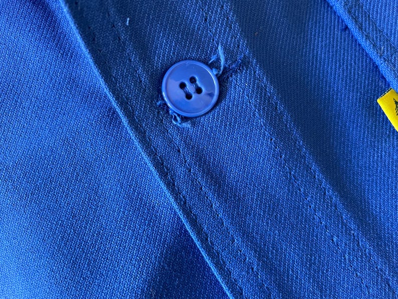 Op de afbeelding: Close-up van een blauwe knoop die op een blauw stoffen shirt is genaaid. De knoop heeft vier gaten.