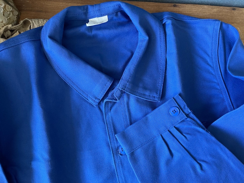 Op de afbeelding: Een blauwe werkjas met een knoopsluiting en een enkele knoop op de mouw.