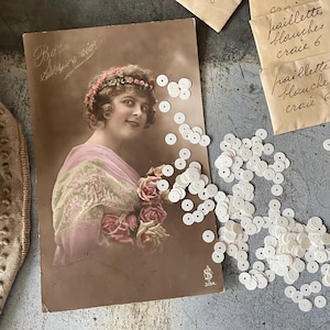 Pode incluir: Uma fotografia antiga de uma mulher usando uma faixa de cabeça floral e um vestido de renda. A foto está em tons sépia e tem um aspecto desbotado. A mulher está segurando um buquê de flores. A foto é cercada por lantejoulas redondas brancas.
