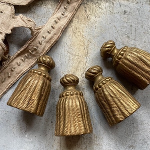 Op de afbeelding: Vier antieke goudkleurige kwasten met een geweven ontwerp en een decoratieve bovenkant. Op de achtergrond een bruin textielafwerking met een gestikt patroon. De kwasten zijn waarschijnlijk voor woondecoratie of knutselprojecten.