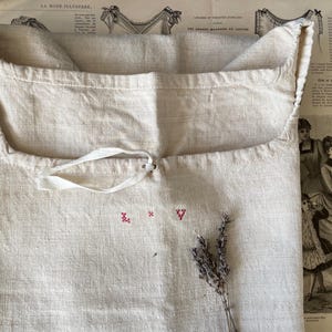 May include: A white linen bag with a drawstring closure and a small embroidered design of the letters "X" and "V" in red thread. The bag is lying on a table with a sprig of dried lavender.