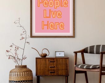 May include: A framed print with the text "Cool People Live Here" in orange letters on a pink background. The print hangs above a wooden cabinet with a small clock and a decorative watering can. A woven vase and a striped chair are also in the room.
