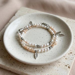 May include: Two pearl bracelets with silver feather charms on a white plate. The bracelets have white and peach pearls. The plate is on a textured, light-coloured surface, creating a soft, neutral aesthetic.