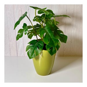 May include: A vibrant green artificial Monstera plant in a light green ceramic pot. The plant features large, glossy, split-leaf foliage. The pot has a smooth, rounded shape, adding a touch of modern decor. The plant is placed on a white surface against a light wood-paneled wall.