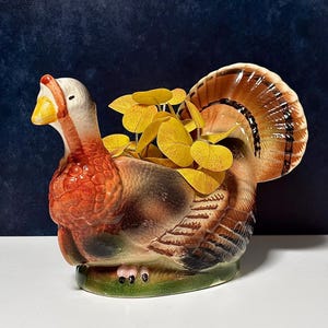 May include: A ceramic turkey-shaped planter featuring a glossy finish with brown, orange, and white coloration. The turkey has a fanned tail and a textured body, with a cluster of artificial yellow leaves emerging from the center opening.