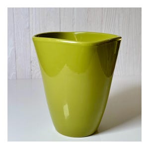 May include: A glossy, lime green ceramic vase with a square top and a tapered body. The vase has a smooth, reflective surface and is set against a white and light wood background. The vase is approximately 8 inches tall.