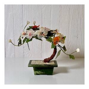 Puede incluir: Un bonsái decorativo con flores blancas, naranjas y rojas. El árbol tiene un tronco marrón y hojas verdes, colocado en una maceta cuadrada verde llena de pequeñas piedras blancas. El fondo es una pared de madera blanca.