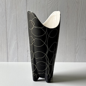 Può includere: Un vaso in ceramica nero con una forma unica e un interno color crema. L'esterno presenta un motivo di linee dorate a forma libera. Il vaso ha una base a tre piedi e un design alto e aperto, adatto per fiori o come elemento decorativo.