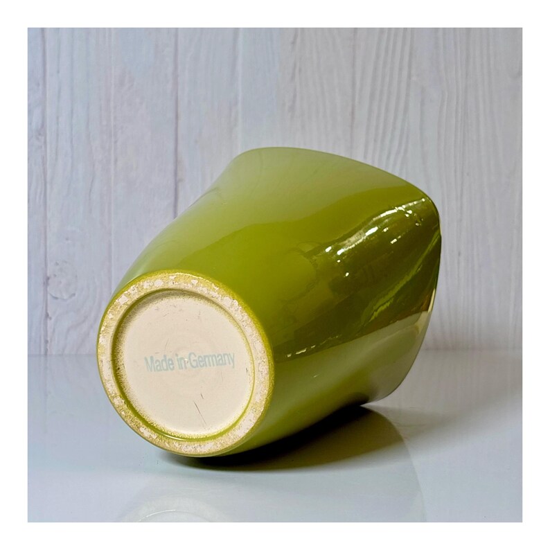 May include: A glossy, lime green ceramic vase with a unique, asymmetrical shape. The bottom of the vase is off-white, with the text "Made in Germany" printed on it. The vase is tilted, showcasing its form and the smooth, reflective surface.