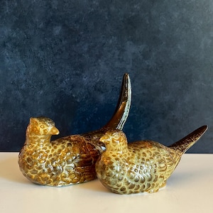 May include: Two ceramic bird figurines with a glossy, mottled brown and tan glaze. The larger bird stands with an upright, elongated tail, while the smaller bird is positioned in front with a shorter tail. Both feature detailed feather textures.