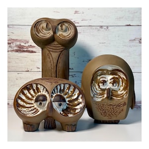 May include: Three ceramic owl figurines in varying shades of brown and cream. One tall, slender owl stands behind two shorter, rounded owl figures. Each piece features hand-painted details around the eyes and textured surfaces on the bodies.