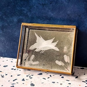 May include: Rectangular mirrored tray with a gold-colored frame. The mirror features an etched design of a bird in flight above cattails. The tray is displayed on a speckled surface against a dark blue background.